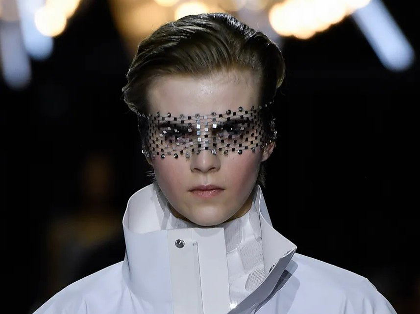 A model on a runway wears a white jacket with a cinched waist, sheer sleeves, and a high collar, paired with a shimmering face veil and fringed