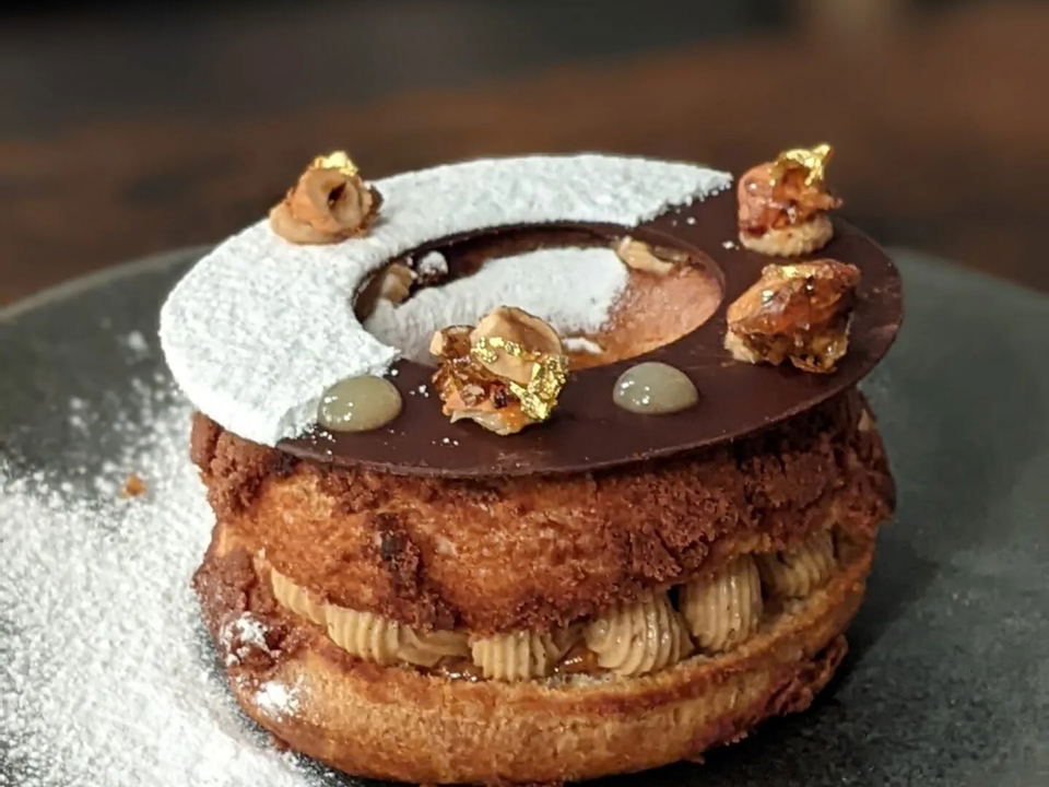 A close-up of a Paris-Brest pastry, a ring of choux dough filled with cream, topped with a chocolate disc, candied nuts, and powdered sugar.