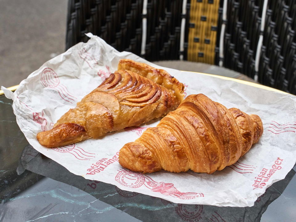 A golden croissant and a caramelized apple tart slice on branded paper, a taste of Paris.