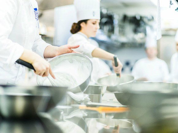 Chefs in white uniforms and toques work in a professional kitchen, mixing ingredients in metal bowls, consistent with culinary school training.