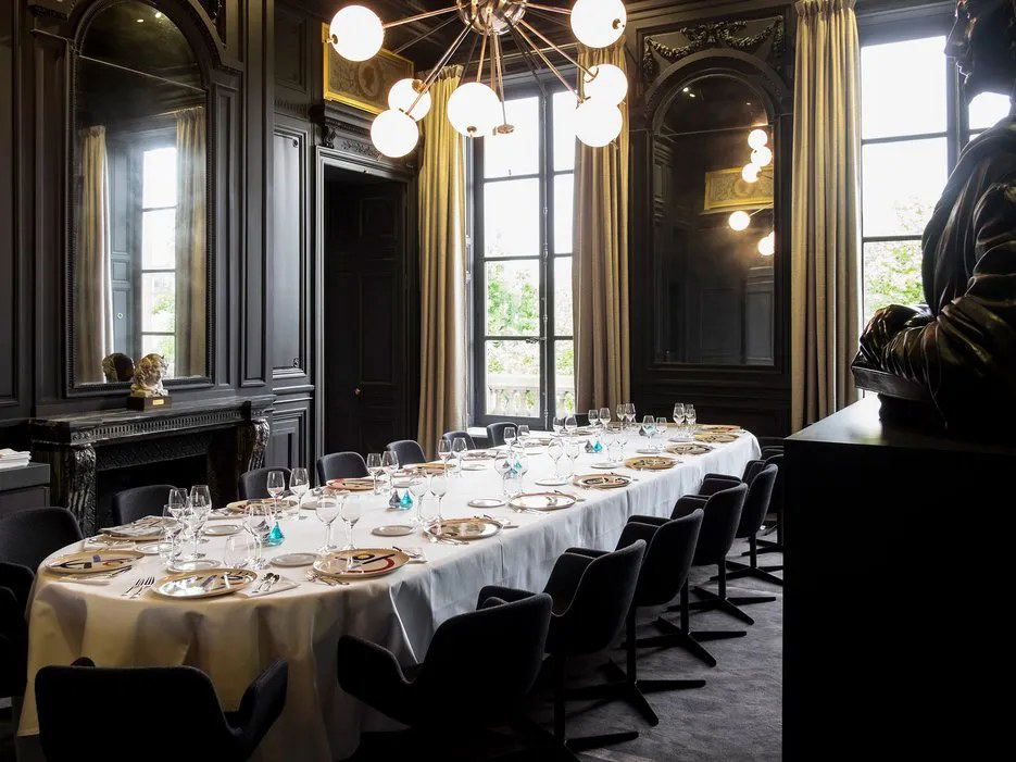 An elegant private dining room with dark paneled walls, a long oval table set with white linen, gold charger plates, and a modern chandelier.
