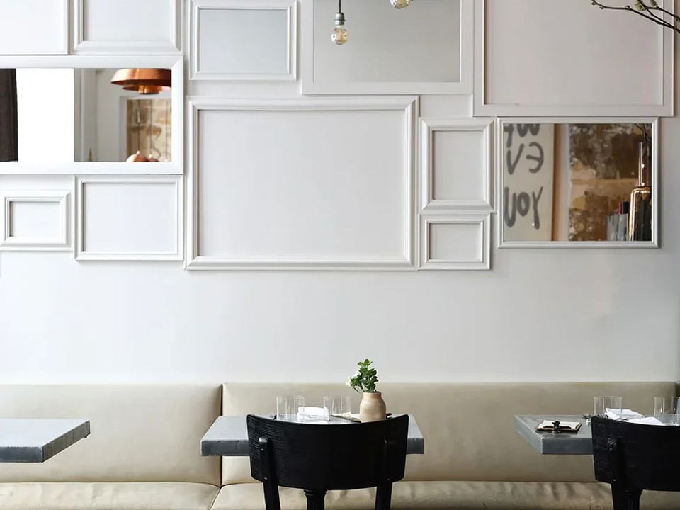 A bright, minimalist restaurant dining room interior with a white wall covered in overlapping white frames and mirrors, cream banquette seating, dark.