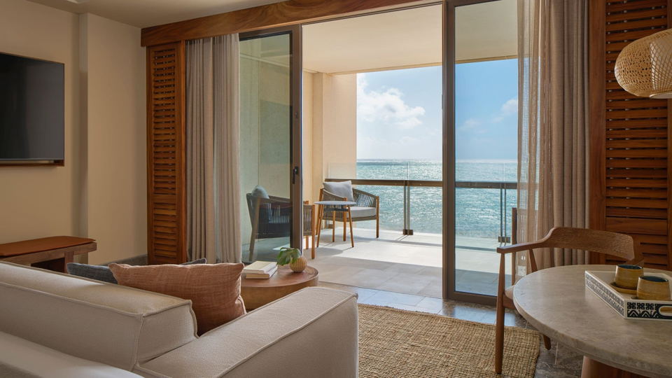 A luxury hotel suite interior with ocean view balcony, featuring contemporary furnishings, neutral tones, wood accents, and floor-to-ceiling glass doo