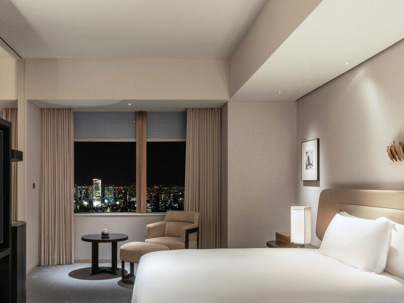 A renovated guest room at Park Hyatt Tokyo at night, featuring a king bed, armchair, side table, and city views from floor-to-ceiling windows.