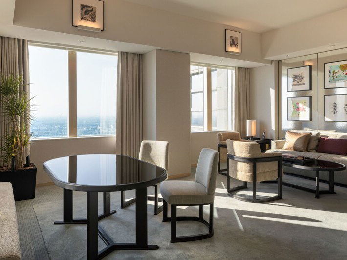 A high-floor hotel suite living/dining area with a dark dining table, upholstered chairs, a sofa, framed artwork, and city-view windows.