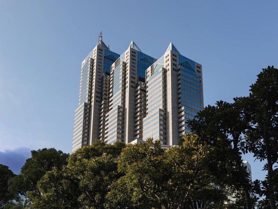 The Shinjuku Park Tower, a tall building with three distinct pointed glass crowns, rises above a canopy of lush green trees against a clear blue sky.