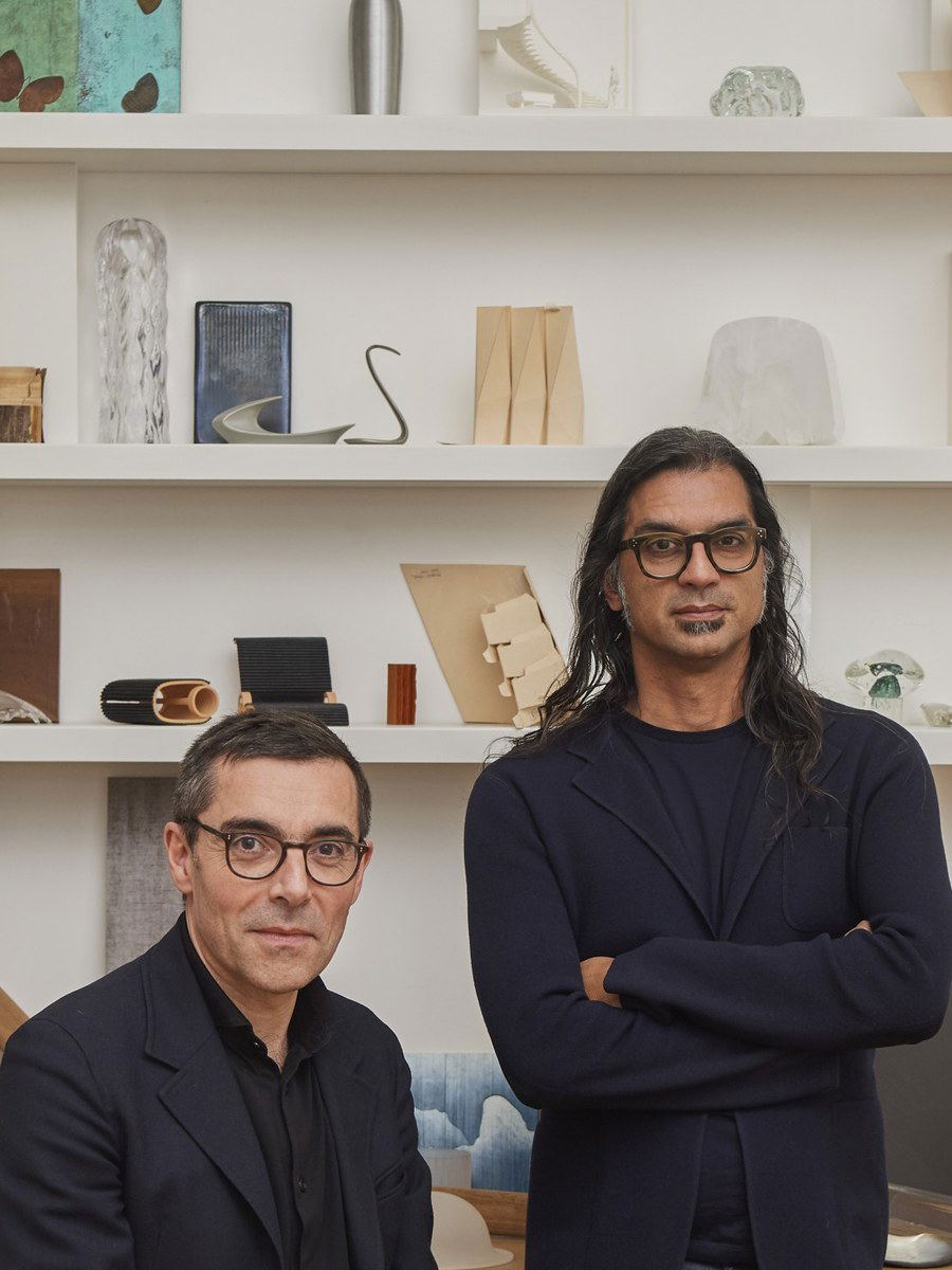 Two men, Patrick Jouin and Sanjit Manku, stand in a studio filled with various design objects on shelves and a desk.