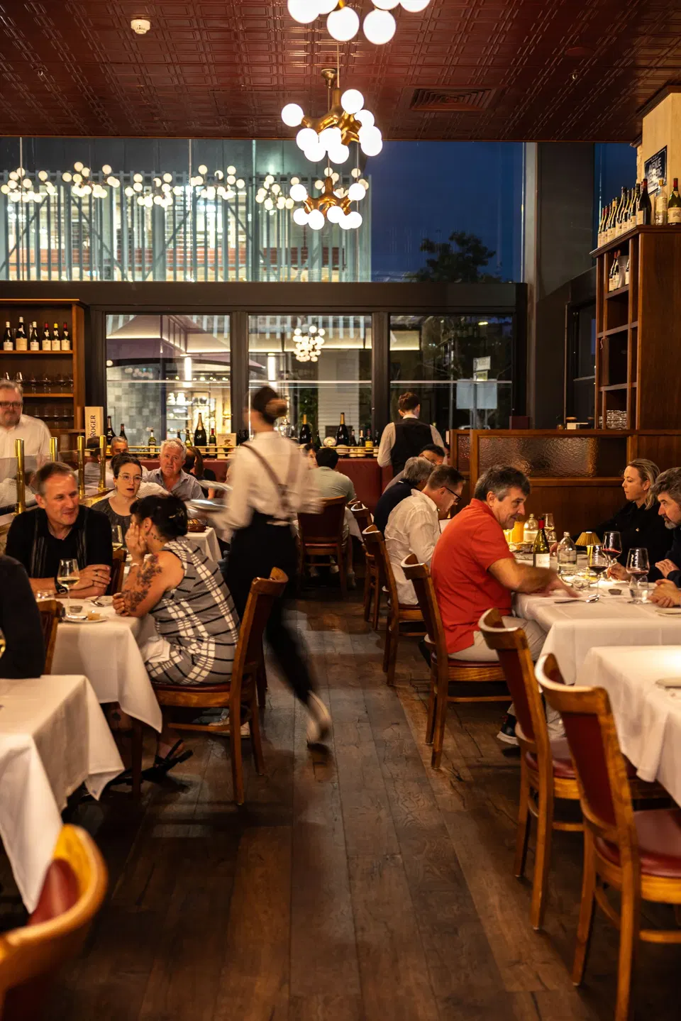 An upscale restaurant dining room with diners seated at white-clothed tables, wooden chairs, decorative globe chandeliers, and large windows showing a twilight cityscape.