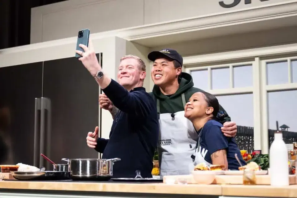 Three participants take a selfie behind pots and plates on a cooking demonstration stage at the 2025 Charleston Classic