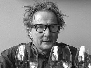 Black and white photograph of a man wearing glasses looking at multiple wine glasses in front of him.