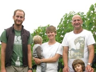 A family of four, two adults and two children, stand in a vineyard with rows of grapevines and ripe dark grapes visible.
