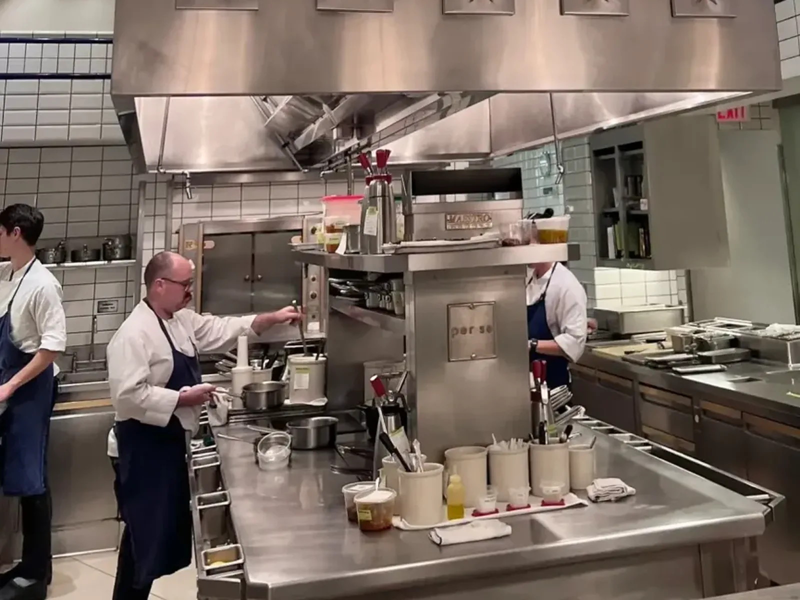 A chef in a white coat and blue apron works at a stainless steel counter in a professional kitchen, surrounded by various cooking utensils and