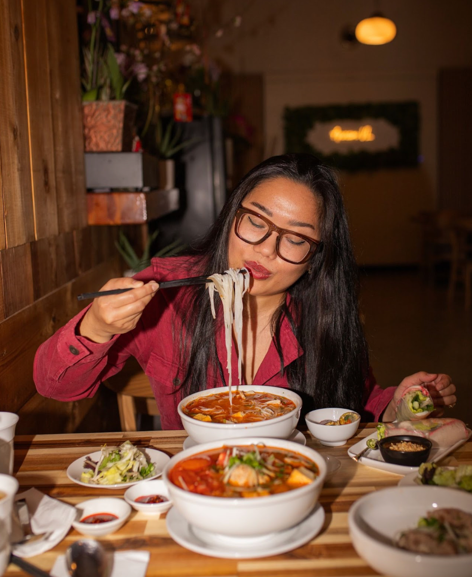 A woman wearing glasses and a red shirt lifts noodles with chopsticks from a bowl of soup at a dining table with several Asian dishes.