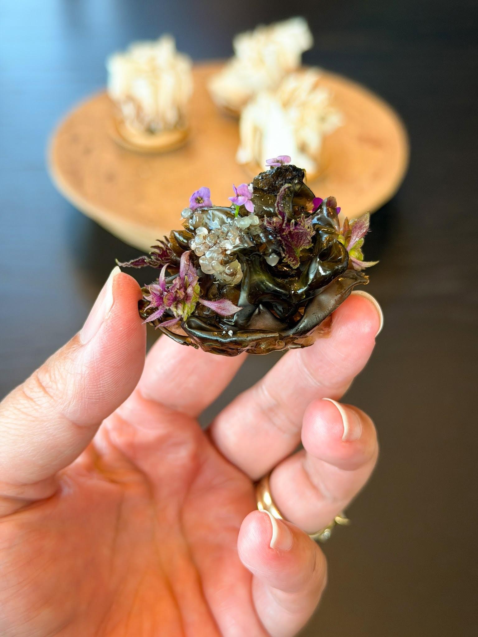 A hand holding a small oyster shell topped with purple flowers, green microgreens, and edible garnishes, with plated desserts blurred in the background.