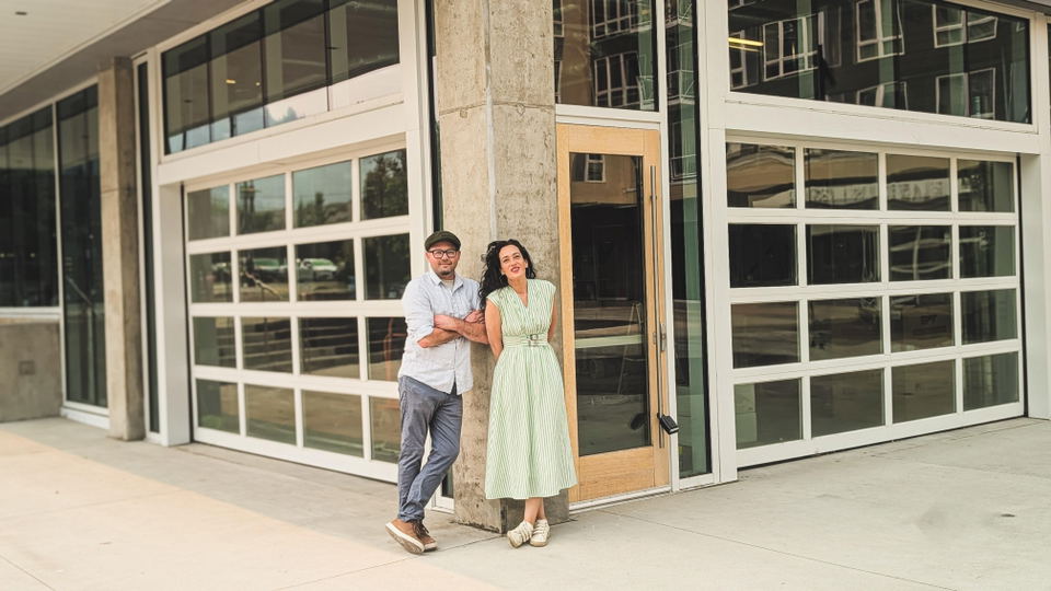 Two people posing back-to-back against a concrete pillar outside a modern building with white-framed glass doors; the source page (Eater Portland) exp