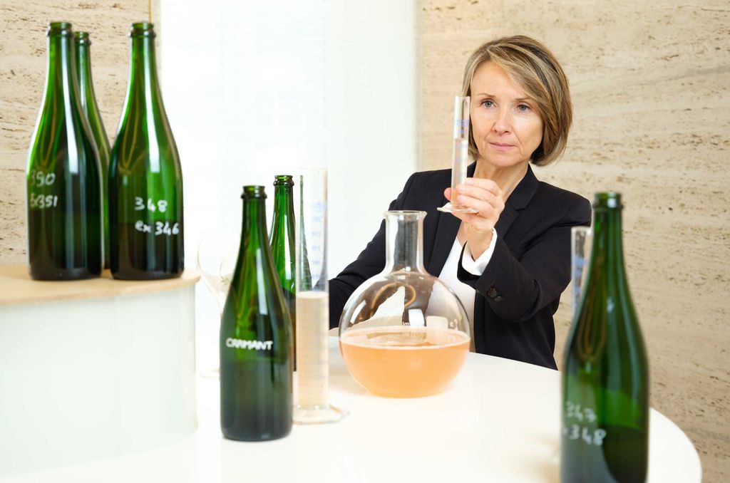 A woman in a black blazer holds up a glass of light-colored wine for inspection, with several green glass bottles arranged on a white table in front of her.