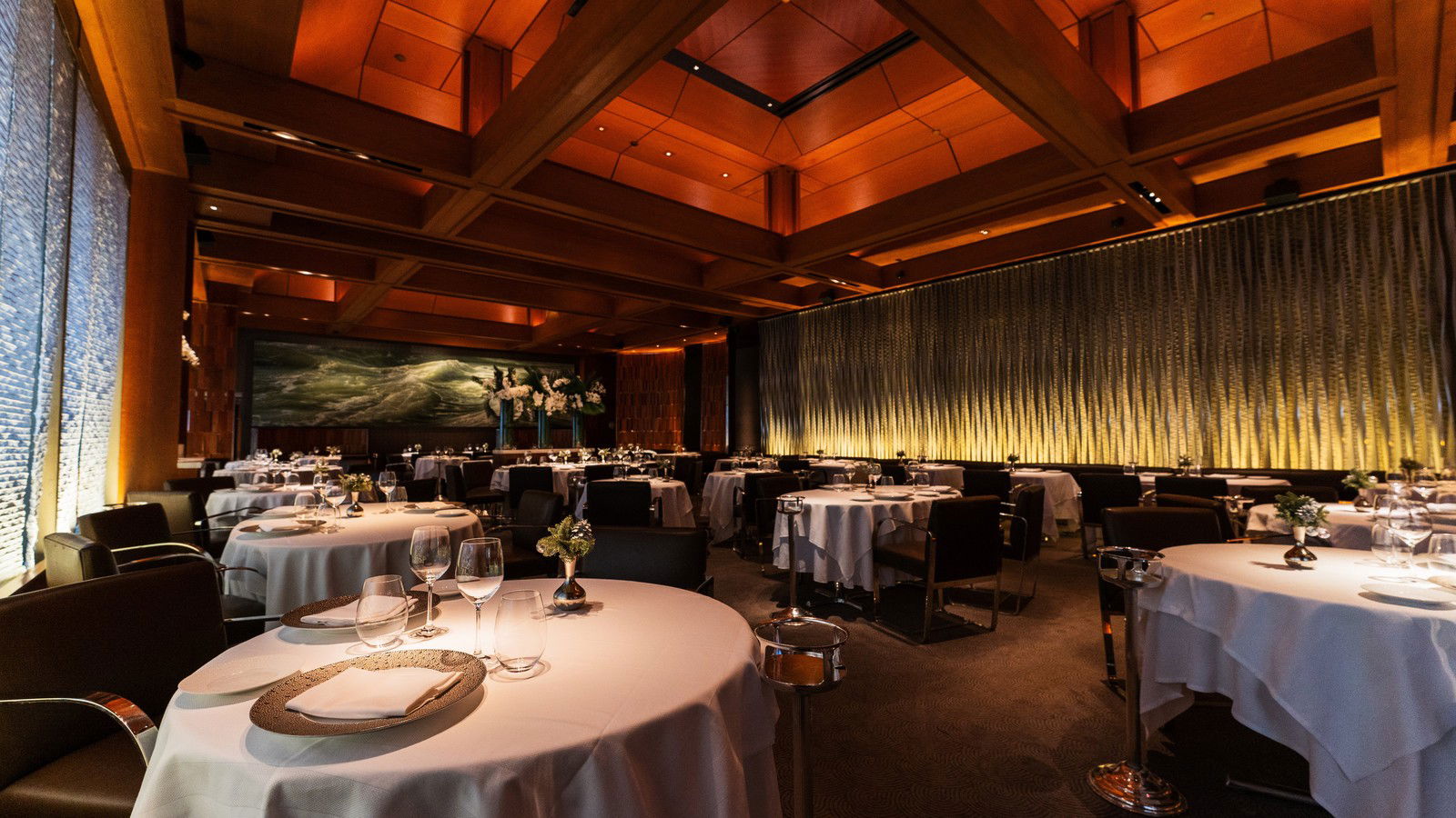 An elegant, warmly lit fine-dining room with white-tablecloth round tables, dark upholstered chairs, and a coffered wood ceiling.
