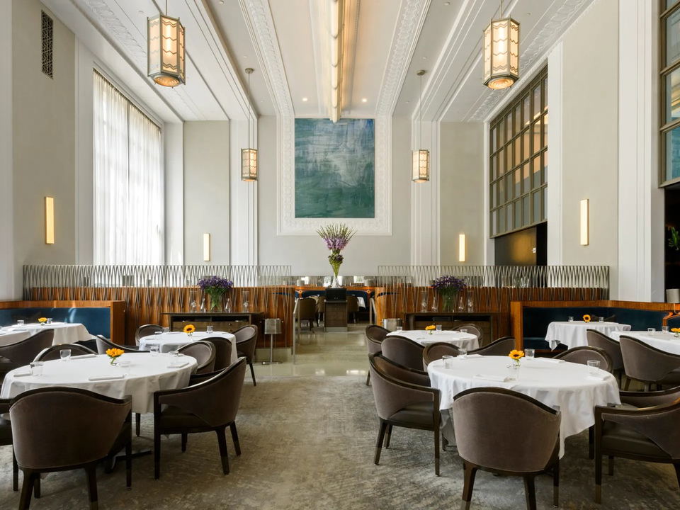 A grand dining room with high ceilings, white tablecloths, brown upholstered chairs, and teal banquettes. Ornate pendant lights hang from the