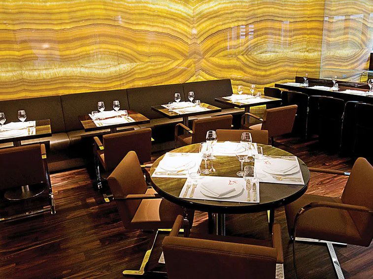An upscale dining room at Marea restaurant features a warm, backlit onyx wall, dark banquette seating, and formally set tables with wine glasses.