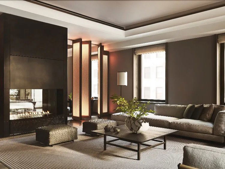 A luxurious hotel suite living room with a double-sided fireplace, neutral upholstered sofas, dark wood paneling, glowing lattice columns, and large