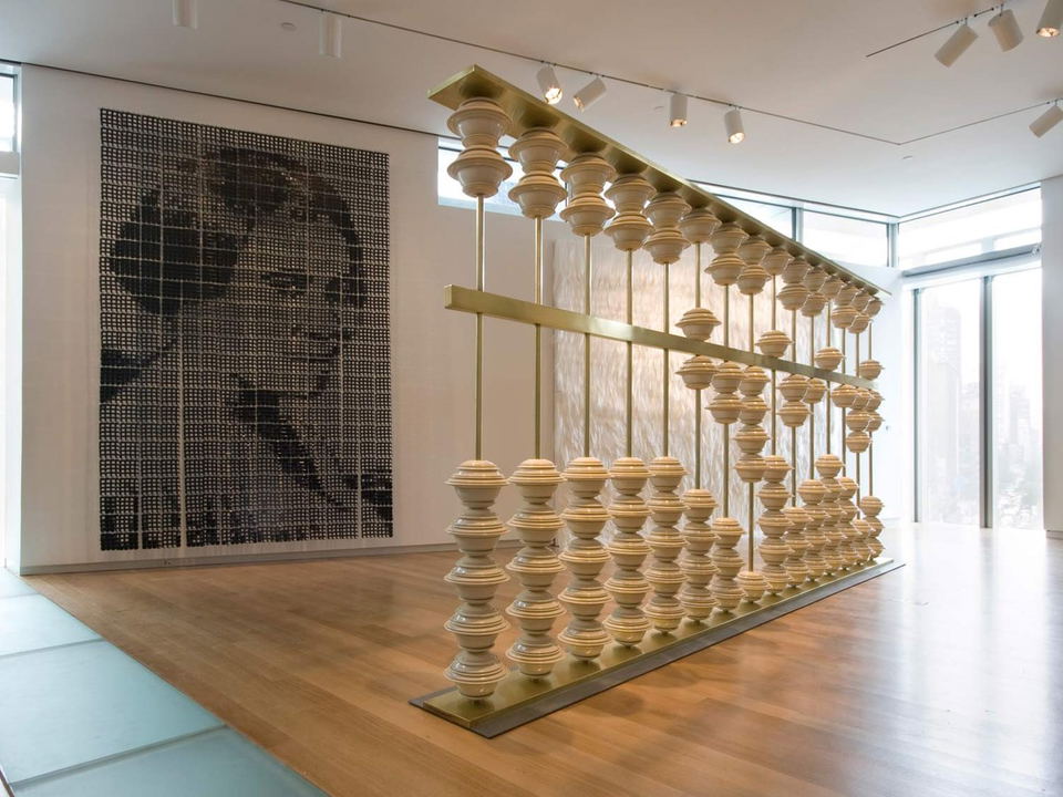 A bright, modern museum gallery features a large-scale sculpture of stacked ceramic forms on brass rods, a pixelated portrait on the wall, and a