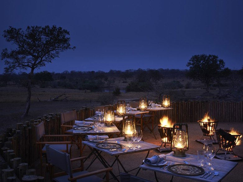 A beautifully set bush dinner scene at night, with lantern-lit tables, glowing fire baskets, and acacia trees silhouetted against a deep blue