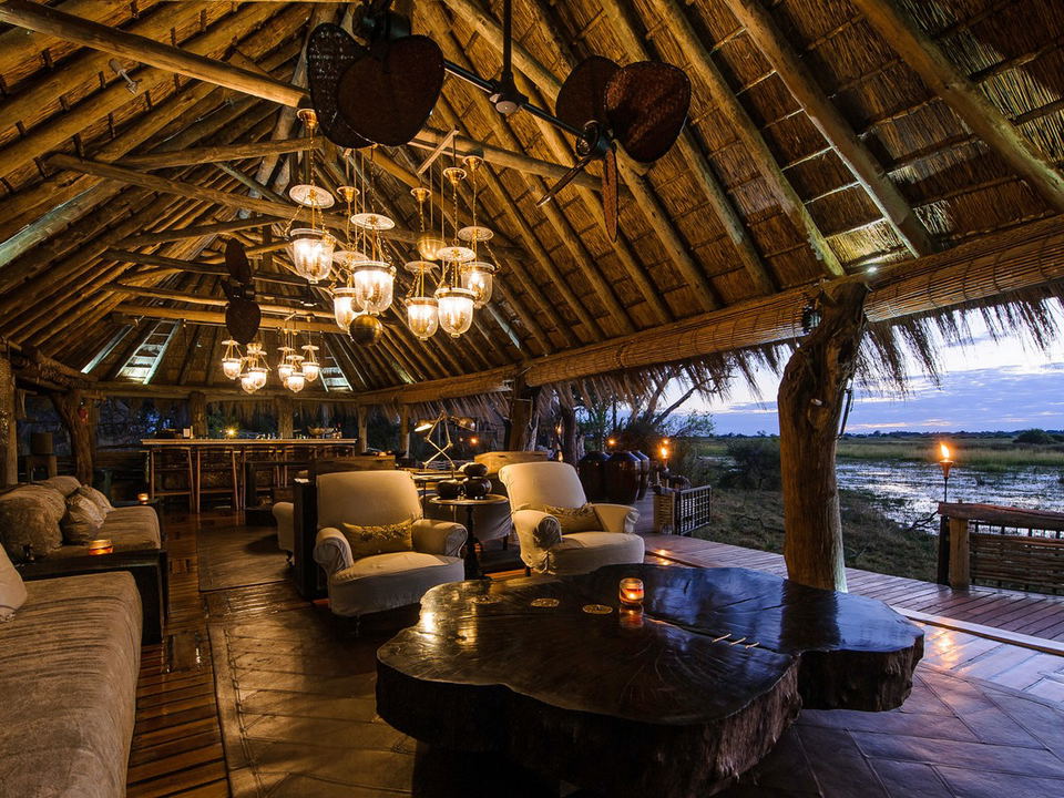 An elegantly lit safari lodge lounge at Mombo Camp, Okavango Delta, with a thatched roof, chandeliers, plush seating, and an open deck overlooking a