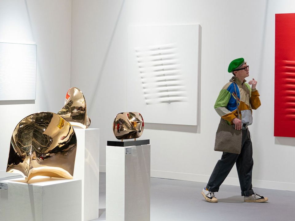 A visitor in a green beret and colorful jacket observes a red geometric relief artwork at the Art Basel Hong Kong art fair, with bronze sculptures