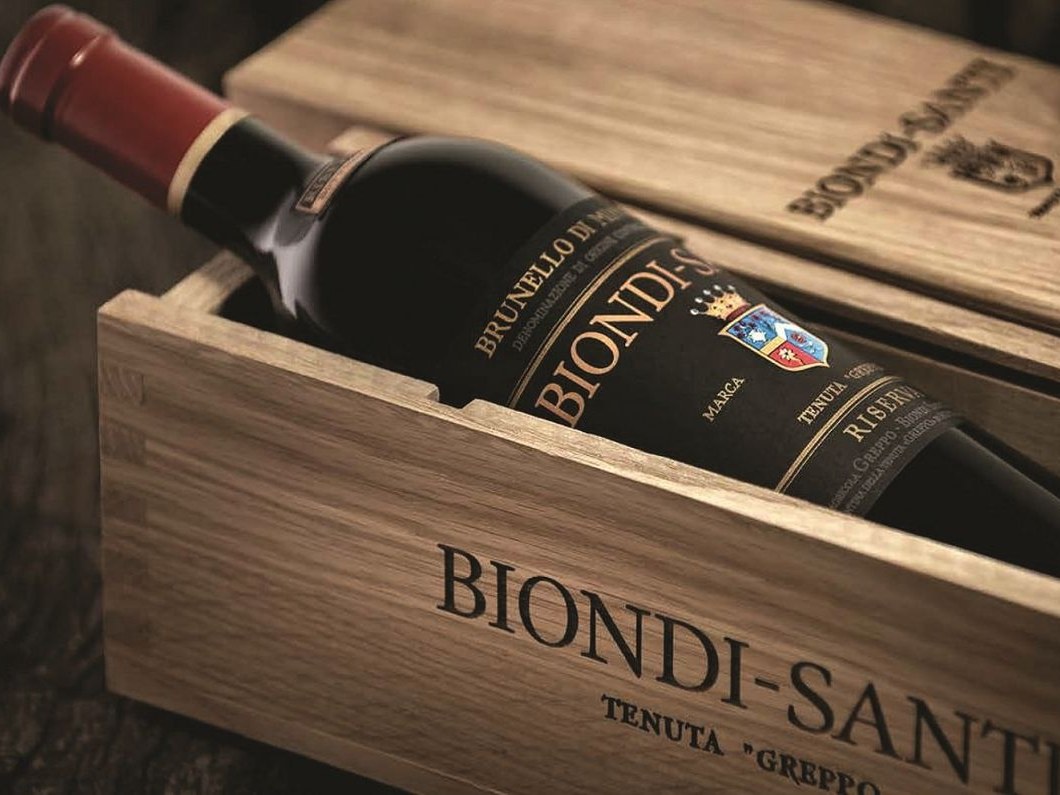 A bottle of Biondi-Santi Brunello di Montalcino Riserva in an open wooden case, showing its black label with gold text and a red foil top.