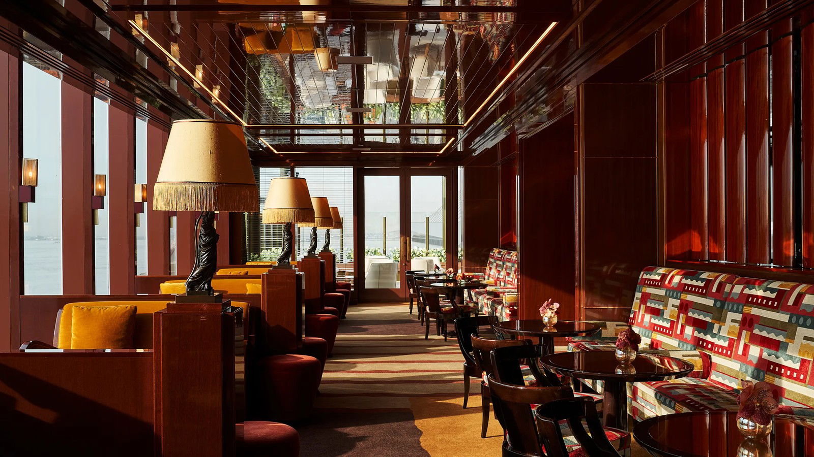 A warmly lit bar or lounge interior with deep mahogany paneling, mirrored ceiling, fringed lamps, and colorful banquette seating.
