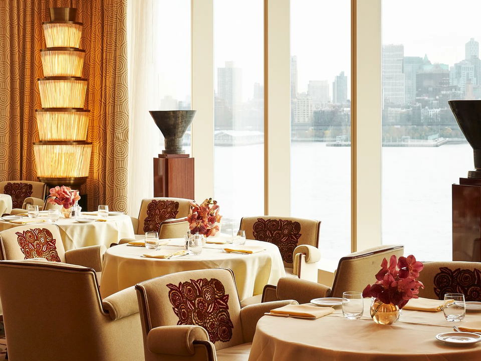 An elegant dining room at Casa Cipriani with round tables, cream chairs with embroidered backs, tiered lamps, and large windows overlooking a city