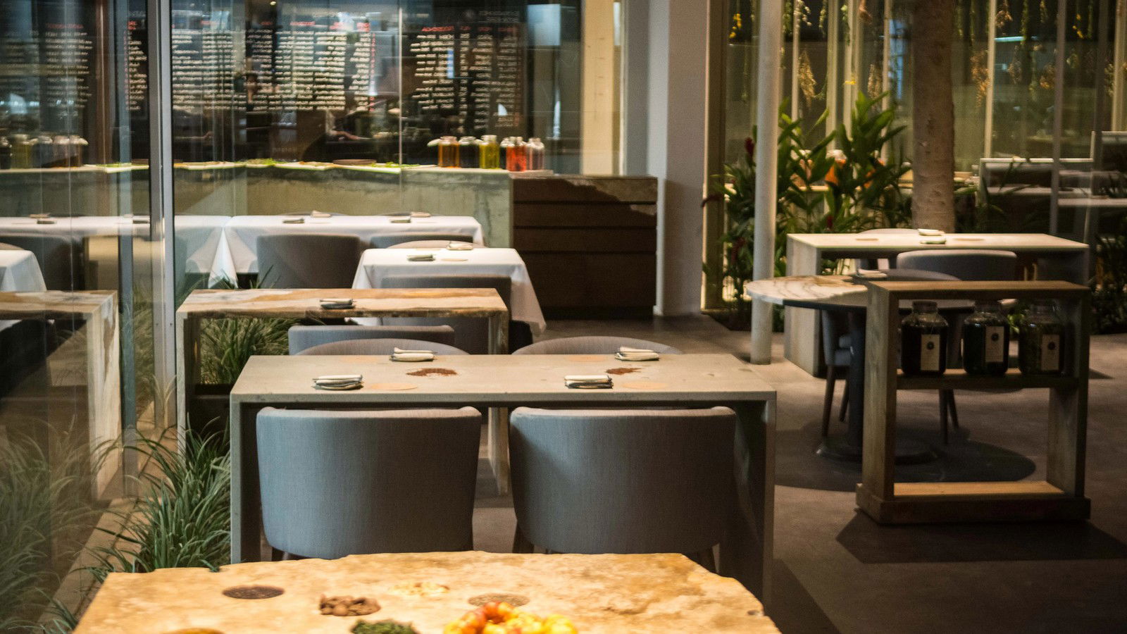 A modern dining room with concrete tables, grey chairs, and white-linened tables in the background, featuring plants and a display table with various