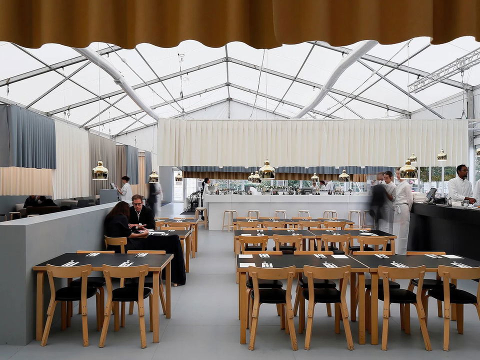 A large, airy dining room in a tent, featuring black-topped tables with wooden chairs, gold pendant lights, and staff at a bar.