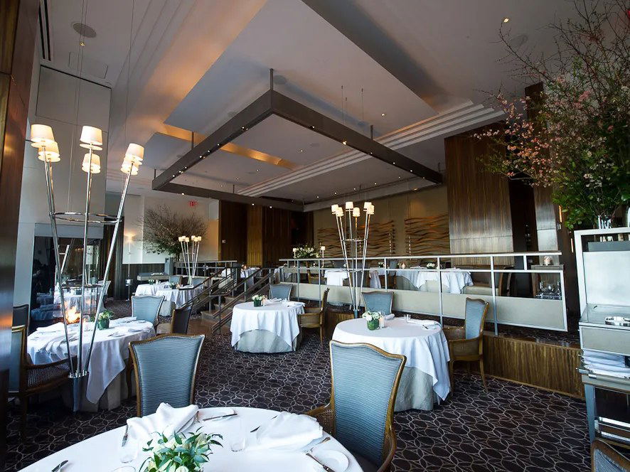 An elegant dining room at Per Se, with white-clothed tables, tall lamps, and cherry blossom arrangements, highlighting an upscale restaurant interior.