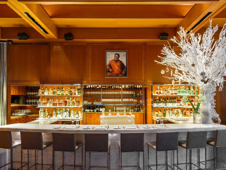 A long, white marble bar counter with several grey bar stools in front of a warm wood-paneled back bar filled with bottles of spirits and glassware.