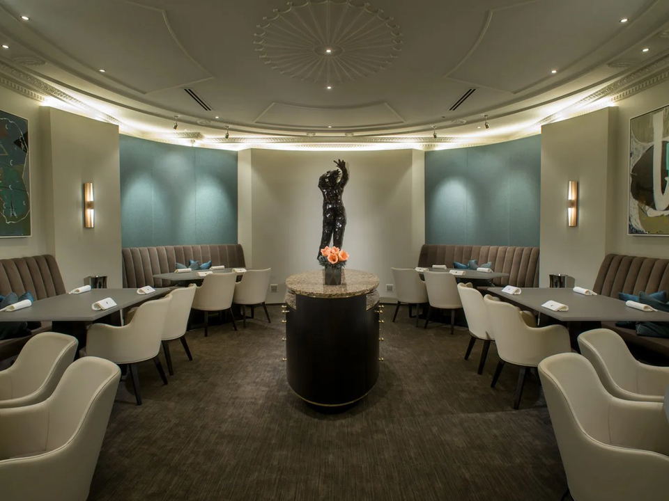 An elegant, oval-shaped fine-dining room with tufted banquette seating, cream armchairs, a central pedestal table with a bronze figurative sculpture.