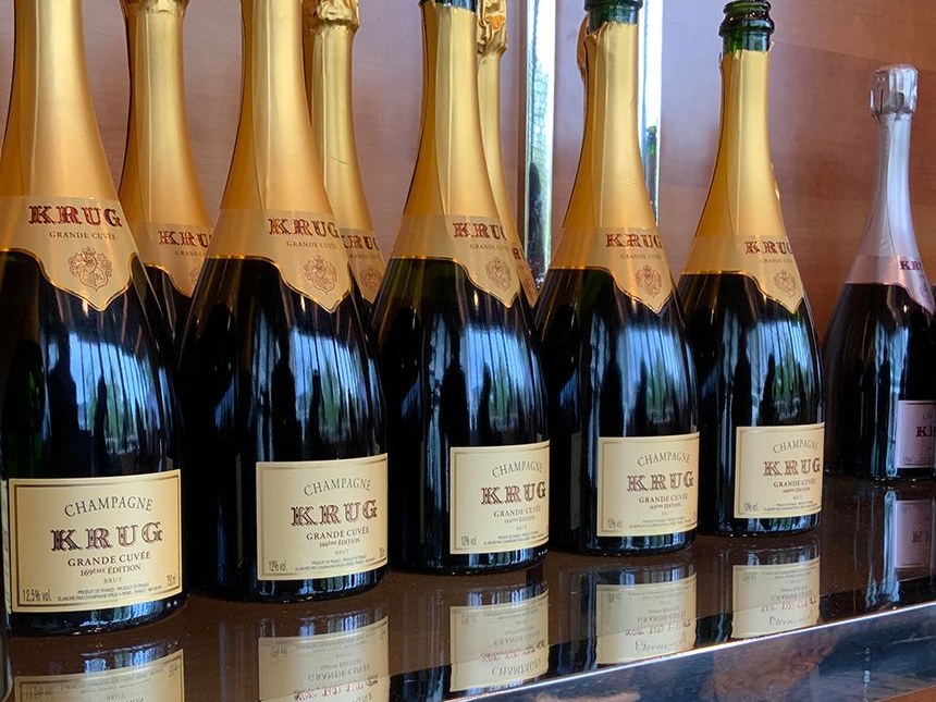 A row of Krug Grande Cuvée bottles, with their distinctive gold foil necks and cream-colored labels, on a reflective surface.