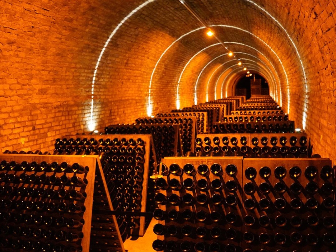 A long, barrel-vaulted brick cellar tunnel in Reims, illuminated by warm amber lighting, filled with thousands of champagne bottles stored on riddling racks.