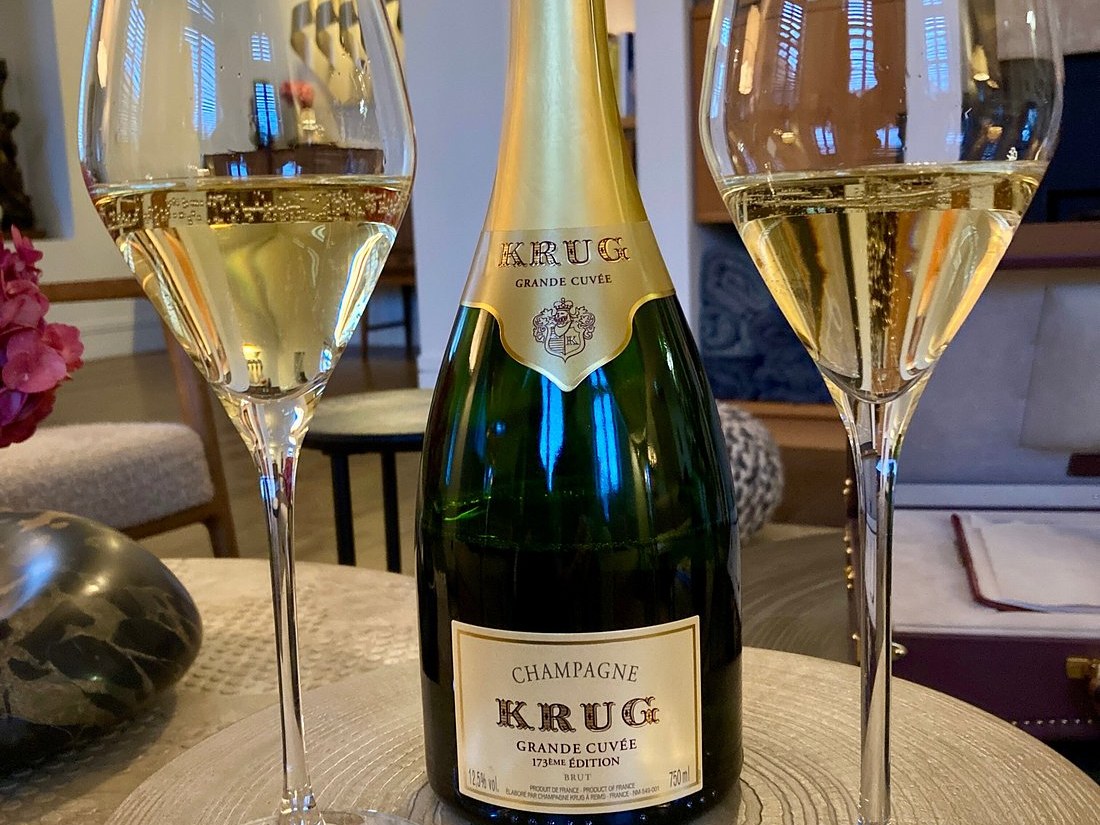 Two champagne flutes filled with bubbly golden liquid flank a dark green bottle of Krug Grande Cuvée 173ème Édition on a light-colored table.