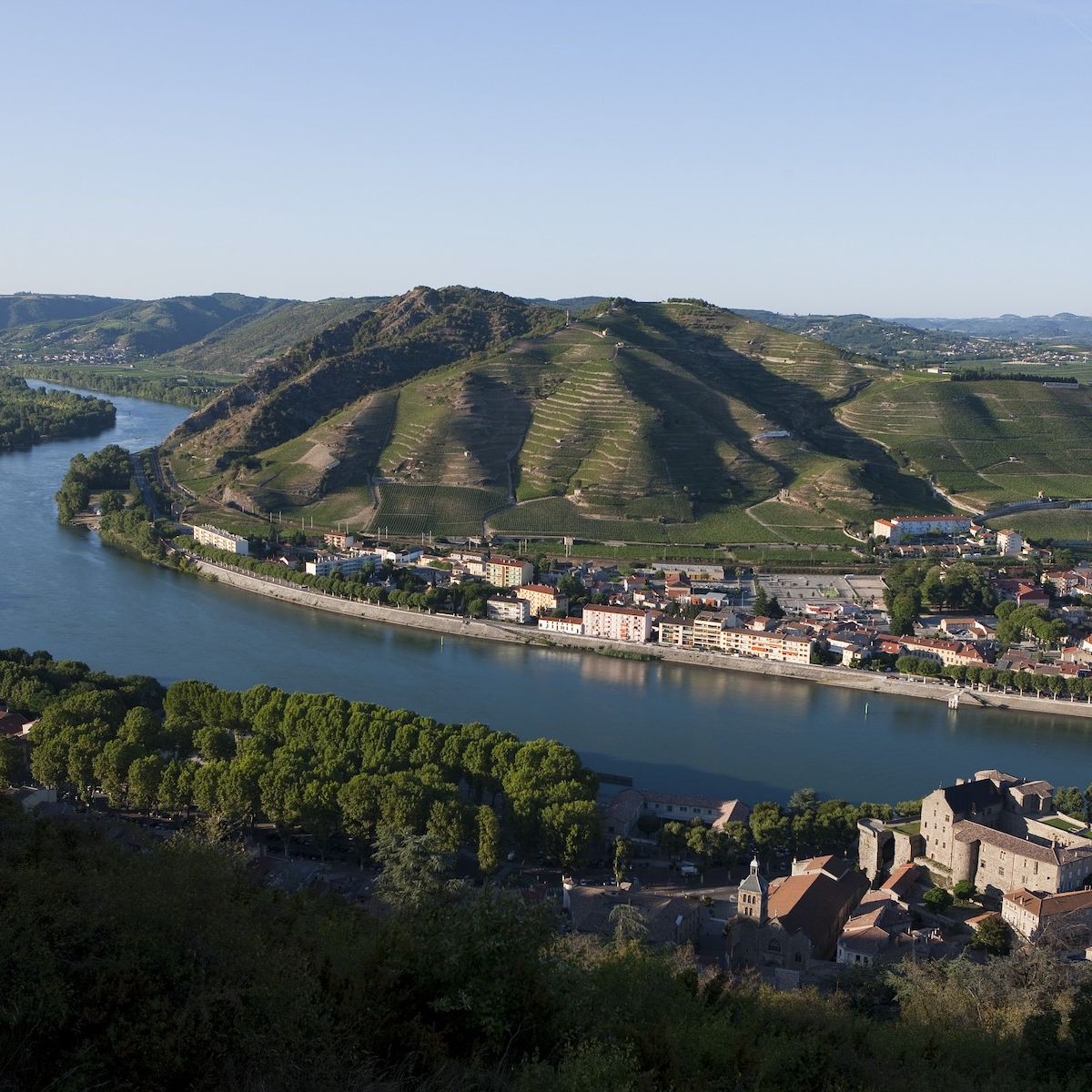 Aerial view of terraced vineyards on hillsides above a riverside town, showing the characteristic granite terraces of the Hermitage hill above Tain-l'.