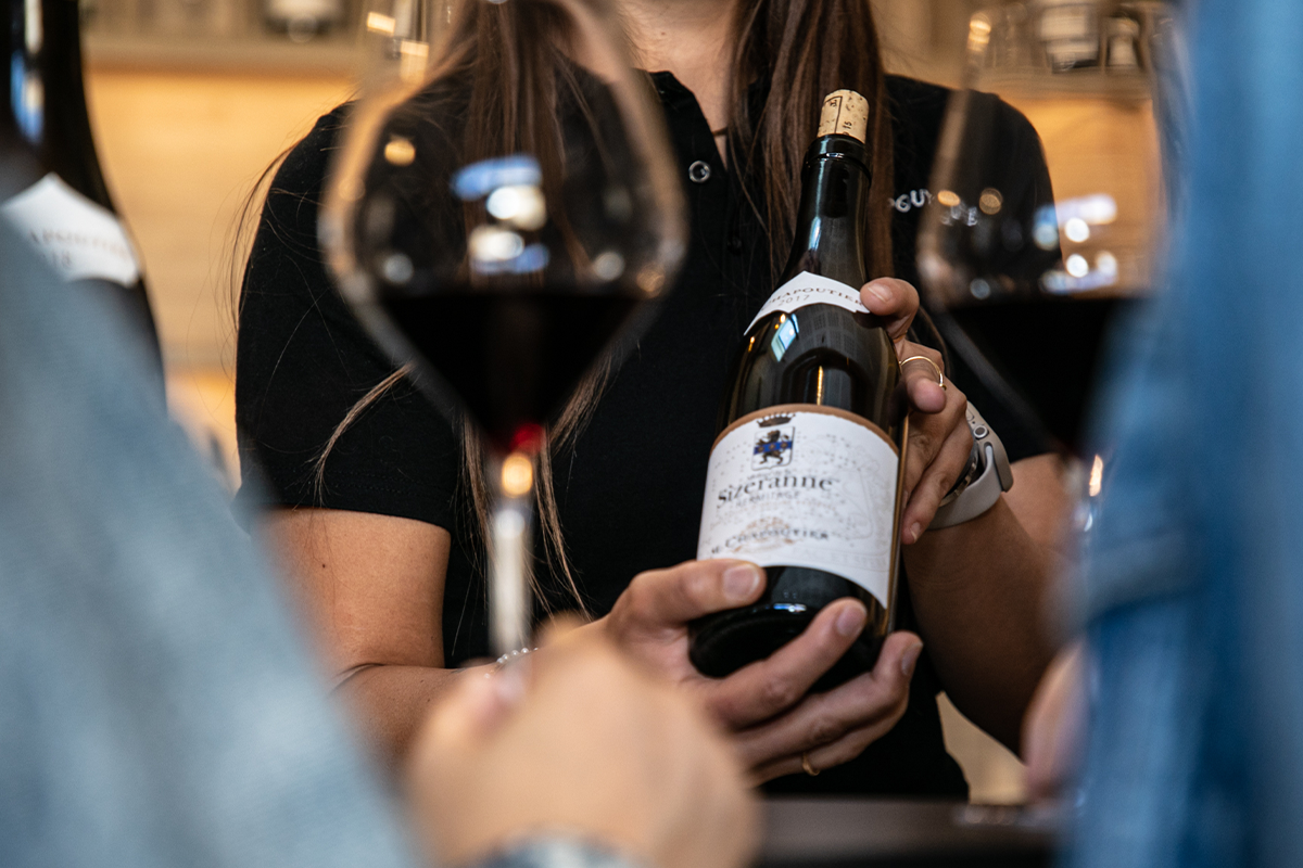 A woman in black clothing holds a bottle of Chapoutier wine while speaking with visitors at what appears to be an indoor event or tasting room.