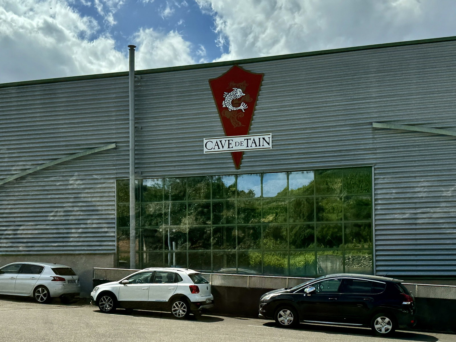 Exterior of Cave de Tain cooperative winery building showing corrugated metal facade with large glass windows, red shield logo with white animal.