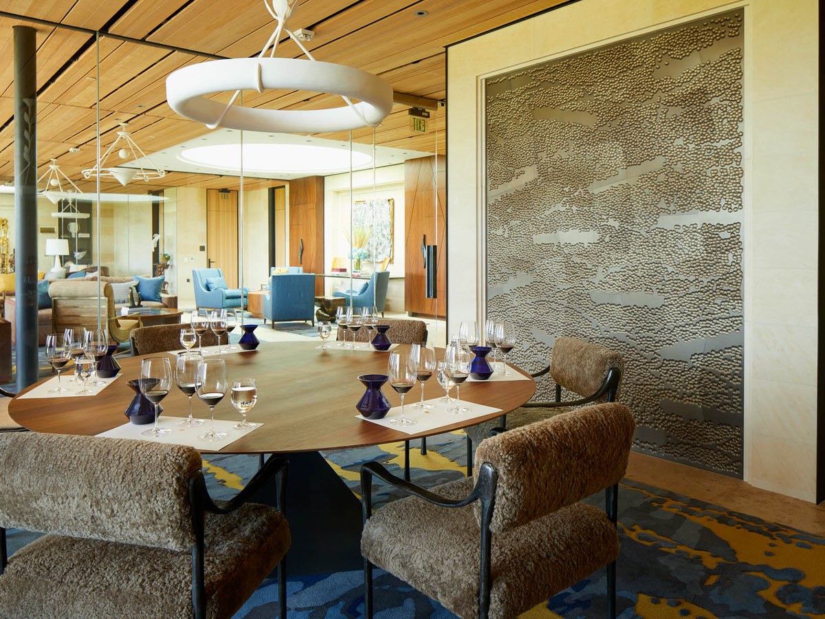 An elegant private tasting room at Opus One, featuring an oval wood table set with wine glasses and blue carafes, surrounded by shearling chairs.