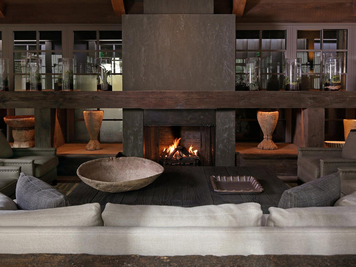 A warmly lit, rustic-luxury hotel lobby interior at Meadowood Napa Valley, featuring a large stone fireplace, dark wood beams, and comfortable