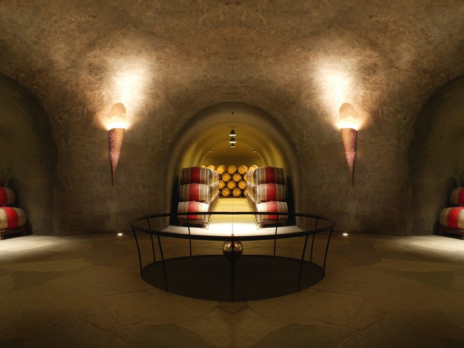 An underground wine cave with arched stone ceilings, illuminated wall sconces, and stacks of oak barrels in a tunnel.