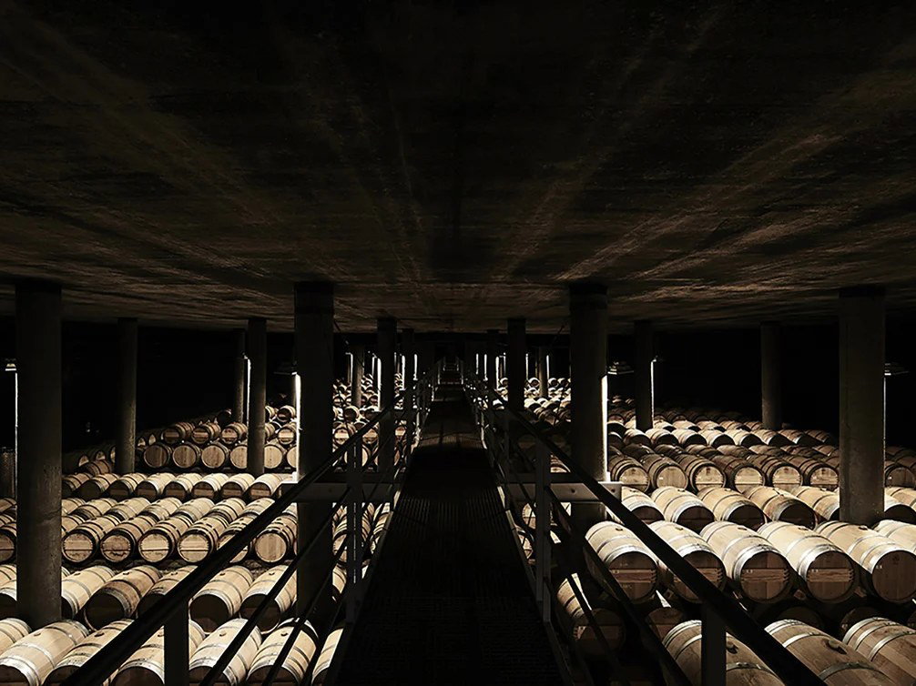 A dimly lit, underground cellar filled with hundreds of wooden wine barrels stacked in long, orderly rows, with a central walkway.