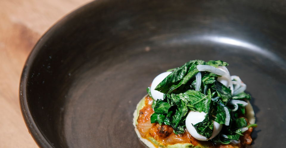 A dish at Pujol featuring a tortilla base, sauce, greens, and onions in a dark ceramic bowl.