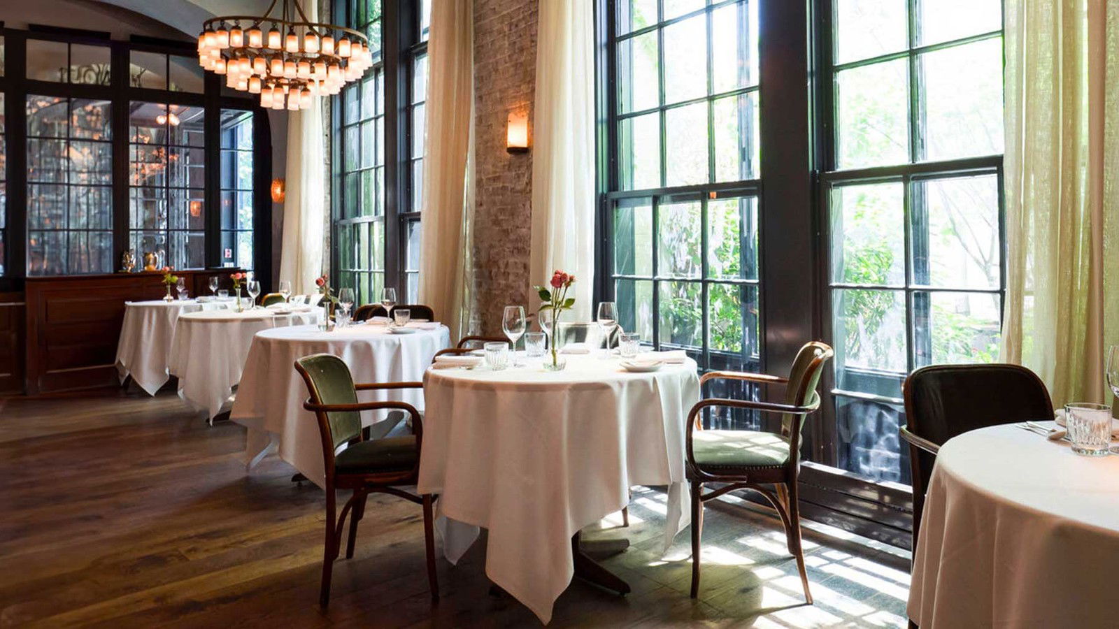 An elegant dining room with white-tableclothed round tables, bentwood chairs, tall windows, exposed brick, and a chandelier.