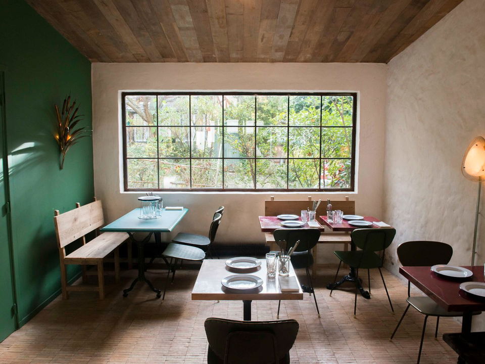 A modern restaurant dining room with wooden ceiling, beige walls, a green accent wall, multiple dining tables with place settings, and a large gridded window overlooking trees.