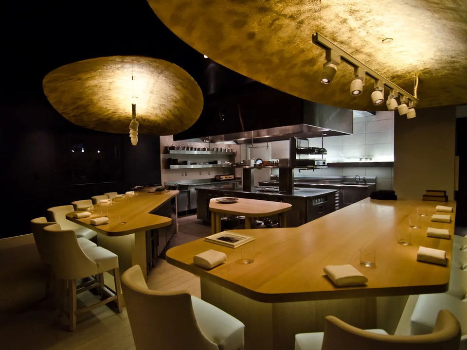 A sleek, intimate restaurant interior with a curved wooden counter and cream upholstered chairs, an open professional kitchen visible behind.
